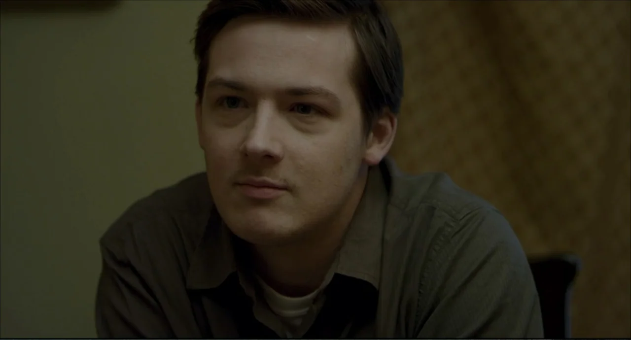 Freedom (2024) movie still: medium — An interior space with a muted olive background and indistinct textured wallpaper. A man in a collared shirt…