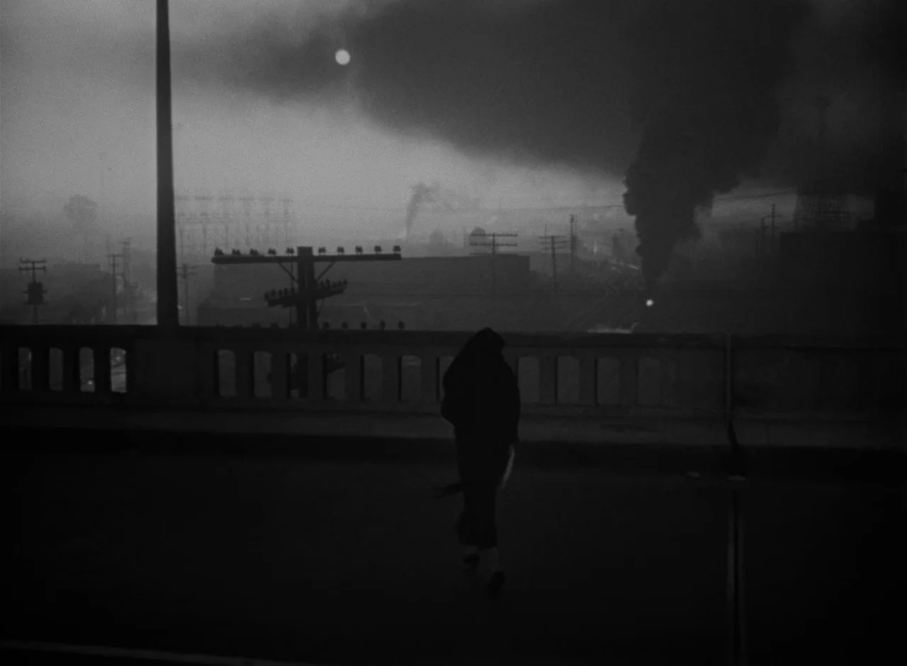 Victims of Sin (1951) movie still: wide — A dark, industrial bridge at night overlooks a smoke-filled urban landscape with power lines. A solitary…