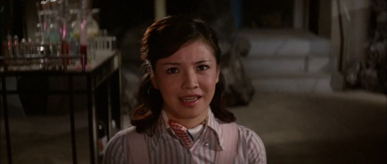 Terror of Mechagodzilla (1975) movie still: medium — A dark laboratory features various glass beakers and test tubes on a metal stand. A woman with…