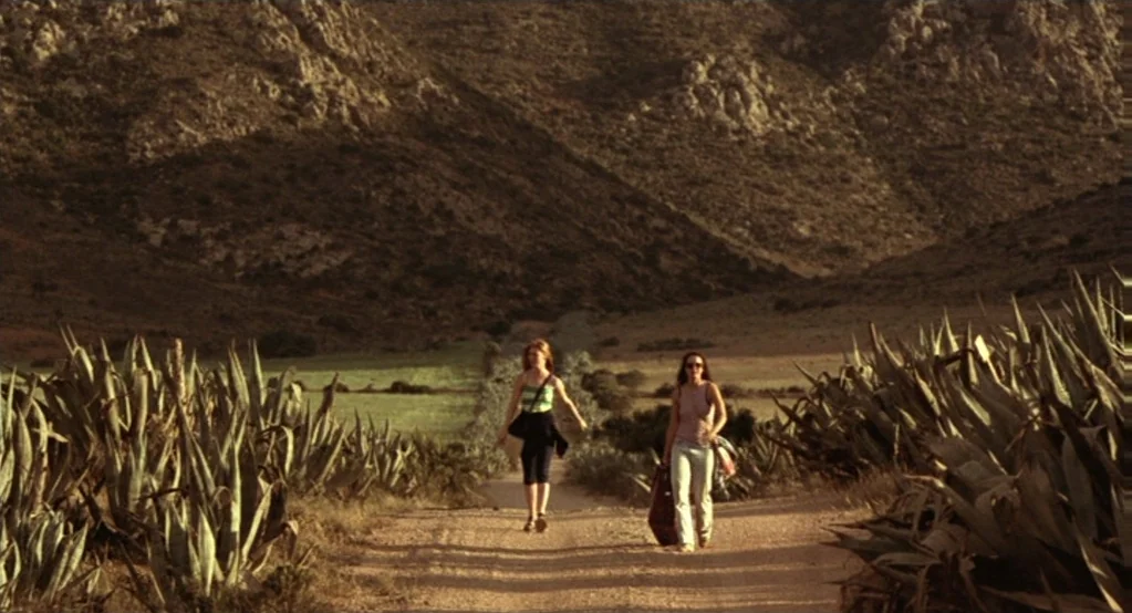 Morvern Callar (2002) movie still: wide — Two women walk down a dirt road flanked by dense agave plants. Large, rocky hills rise in the background…