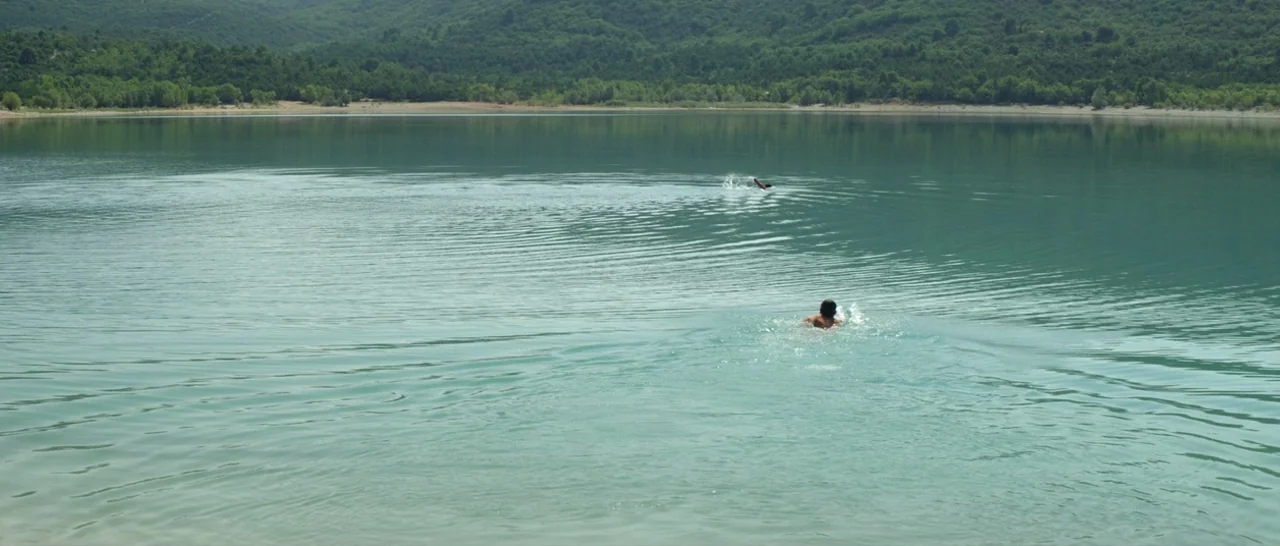 Stranger By The Lake (2013) movie still: wide — A wide lake sits at the base of a forested green mountain slope. Two people are swimming in the open…