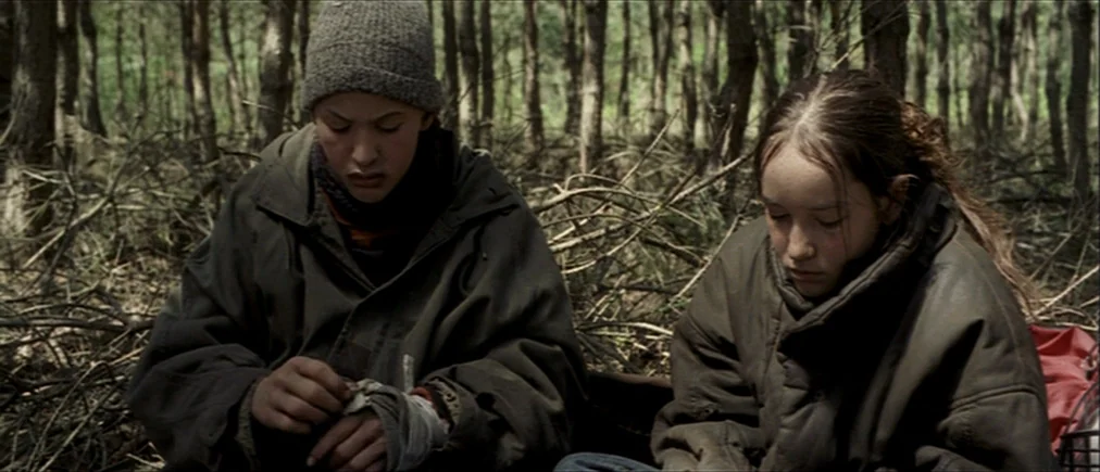 Time Of The Wolf (2003) movie still: medium — Dense forest floor covered in dead twigs and brown leaves under a thick canopy. Two teenagers in heavy…