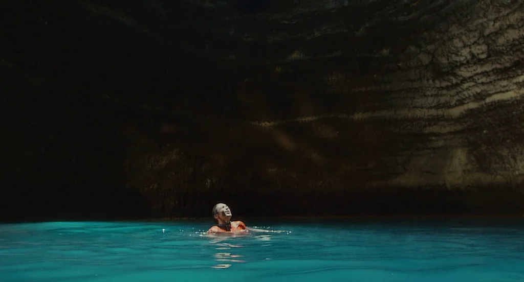 127 Hours (2010) movie still: wide — A deep subterranean pool is enclosed by high, rough-textured cavern walls of dark rock. A single person swims…