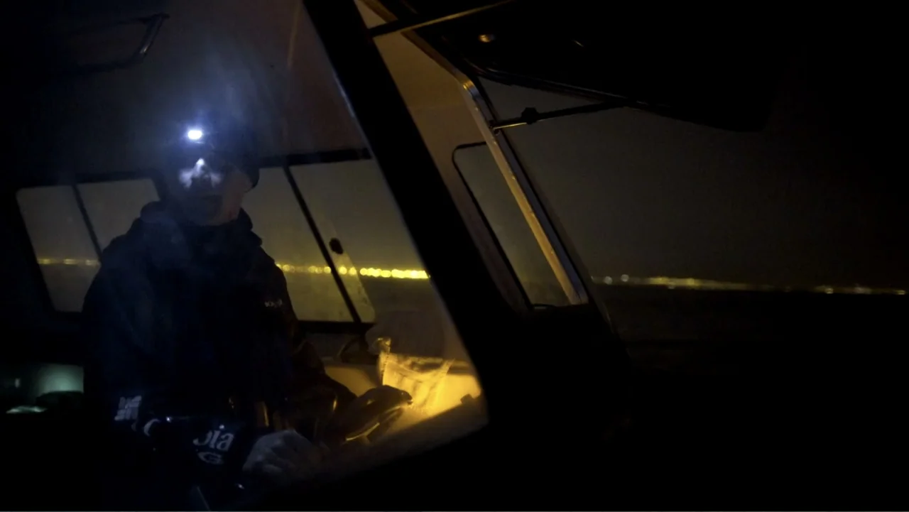 Air Jaws: Night Stalker (2016) movie still: medium — A dark boat cabin at night is illuminated by a small headlamp and city lights. A man sits at…