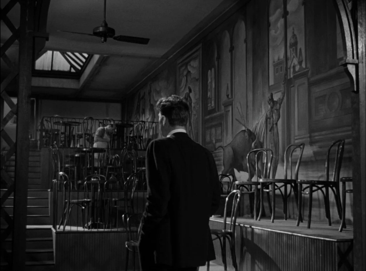 Criss Cross (1949) movie still: medium — An interior cafe space features a large mural painted on the wall. A man in a dark suit stands looking…