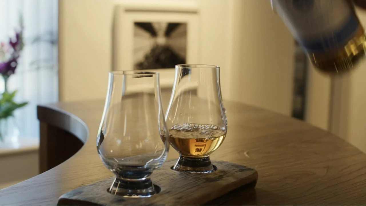 A Scottish Love Scheme (2024) movie still: medium — A close view of two crystal whiskey glasses sitting on a stained wooden serving paddle. A hand…