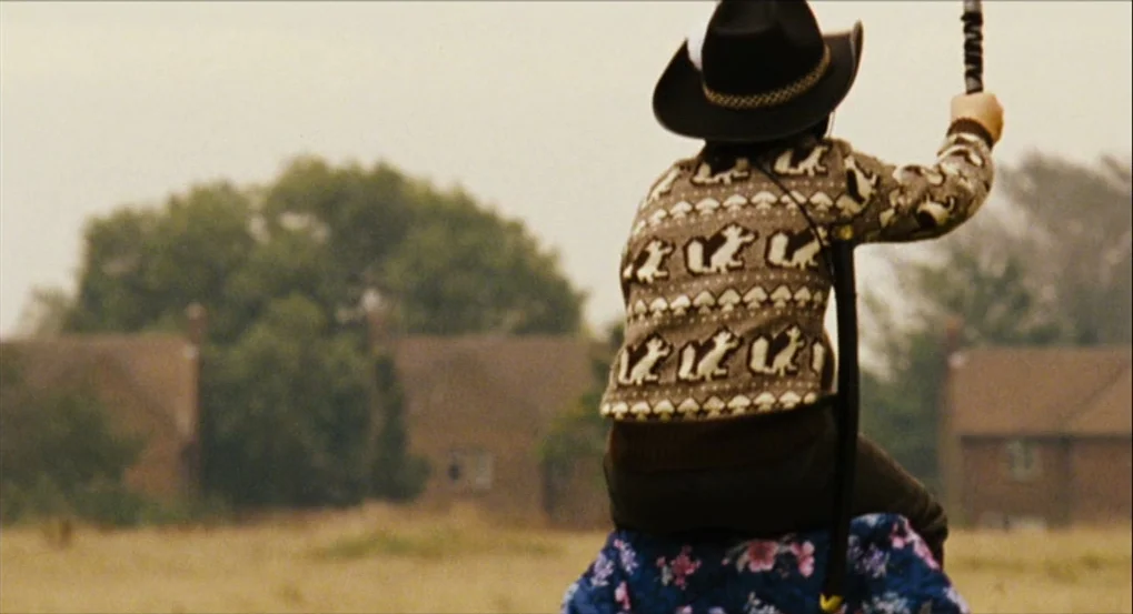 This Is England (2006) movie still: medium — A dry, grassy field sits in front of brick residential houses under a sky. A child in a patterned…