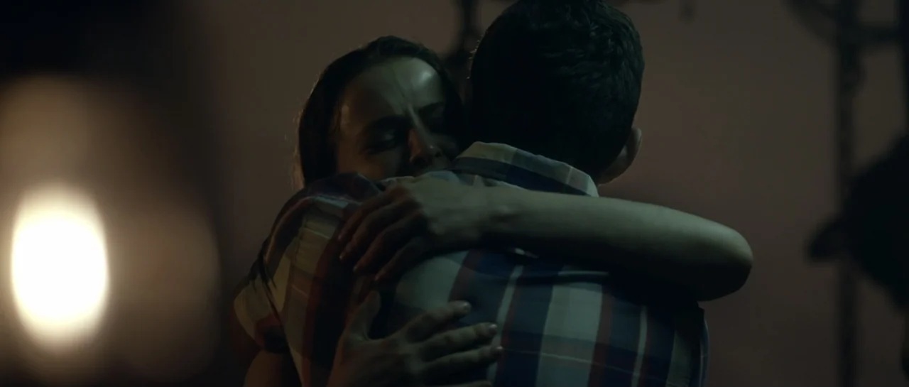 The Lullaby (2019) movie still: closeup — A dimly lit room with a man and woman embracing each other closely. The woman faces the camera with eyes…
