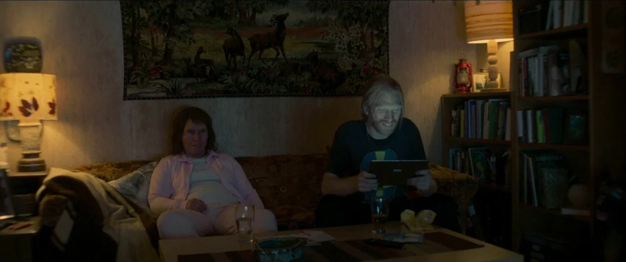 Border movie still: medium — A cluttered living room features a patterned deer tapestry hanging on the wood-paneled wall. A man and woman sit…