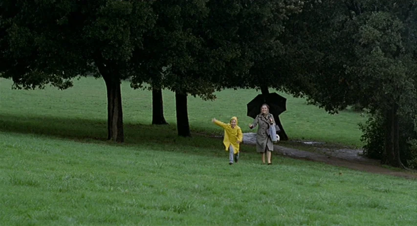 The Fifth Cord (1971) movie still: wide — A rain-dampened grassy hillside is bordered by a line of mature leafy trees. A young child in a yellow…