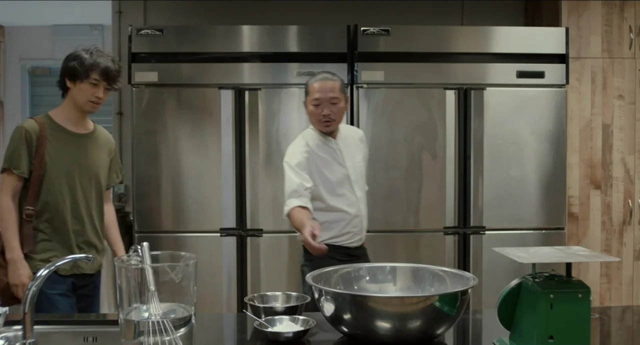 Ramen Shop (2018) movie still: medium — A professional kitchen features stainless steel refrigerators and countertops with various metal bowls. A…