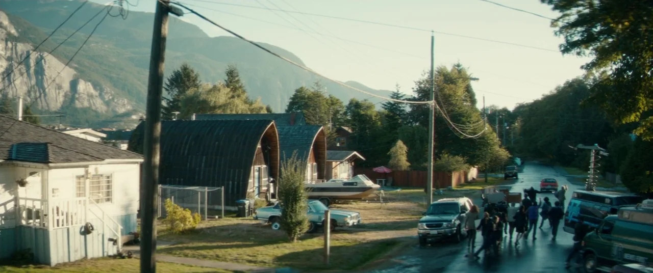 Horns (2013) movie still: wide — A suburban street in a mountainous valley is lined with small cottages and trees. A group of people walks down the…