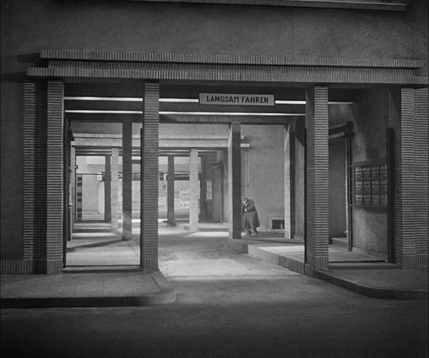 M (1931) movie still: wide — A modernist concrete entrance structure features a sign reading LANGSAM FAHREN over the passage. A solitary older man…