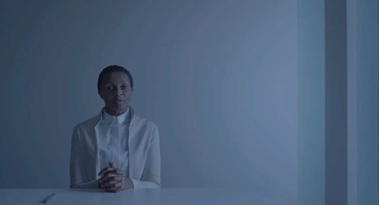 Equals (2015) movie still: medium — A person sits before a plain white wall inside a minimalist room. The subject rests their hands on a white…