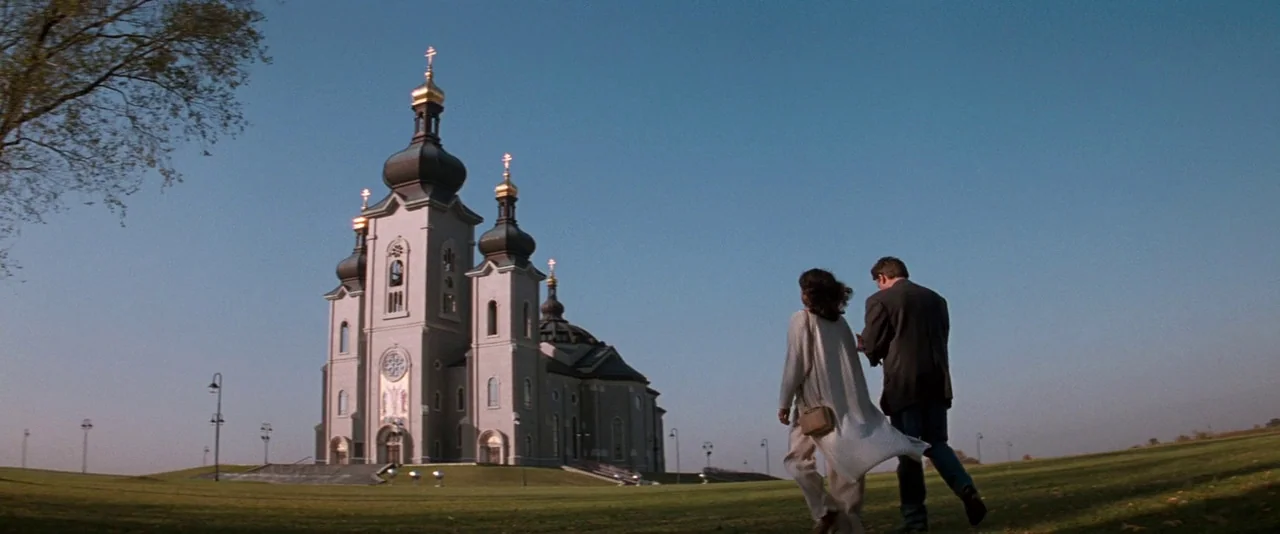 In The Mouth of Madness (1994) movie still: wide — A large stone church with multiple black domes stands on a grassy hill. A man and woman walk away…