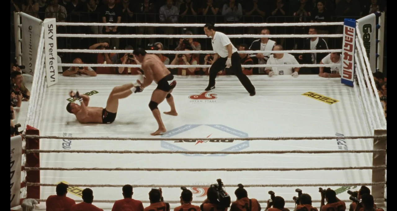 The Smashing Machine (2025) movie still: wide — A professional mixed martial arts fight takes place inside a square canvas ring. Two fighters are in…