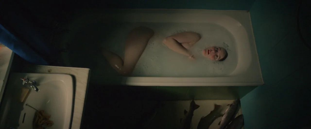Kissing Candice (2017) movie still: wide — A dimly lit bathroom features blue tiled walls and a full bathtub of water. A young woman lies in the…