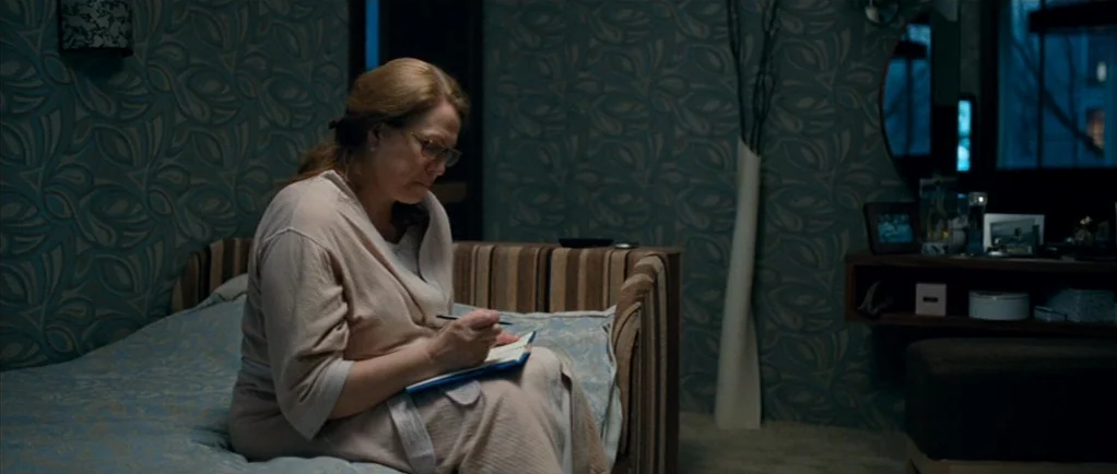 Elena (2011) movie still: medium — An interior hotel room features patterned blue wallpaper and a bed with blue bedding. An elderly woman sits on…