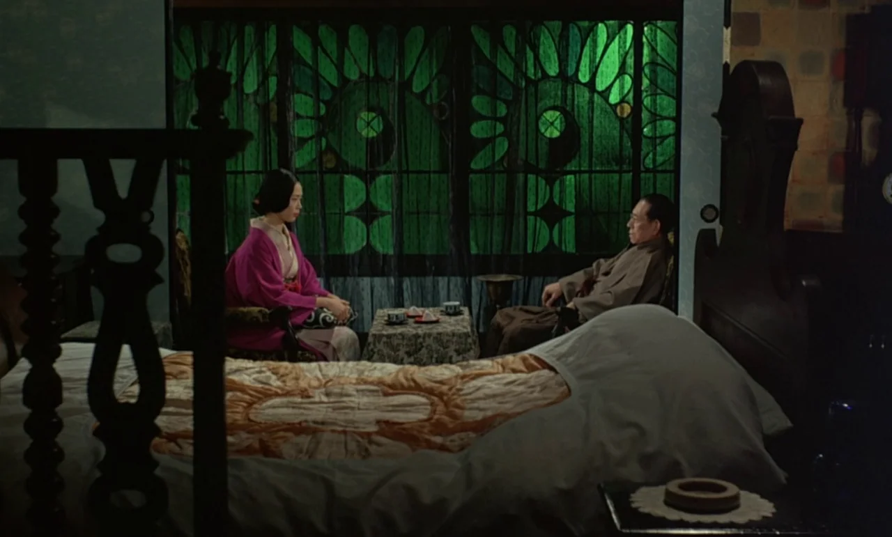 In The Realm of the Senses (1976) movie still: wide — A dimly lit room features a pair of subjects sitting by an ornate window. A large, rumpled bed…