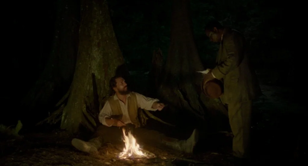 Free State of Jones (2016) movie still: fullbody — A dark swamp area at night features two large tree trunks with flared roots. One man sits by a…