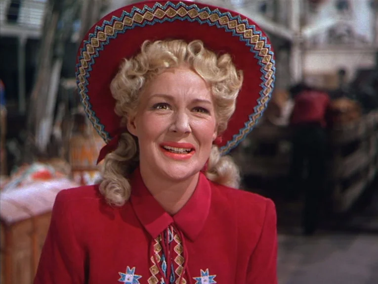 Annie Get Your Gun (1950) movie still: closeup — A woman sits inside a train car with wooden paneling and indistinct baggage. She wears a festive…