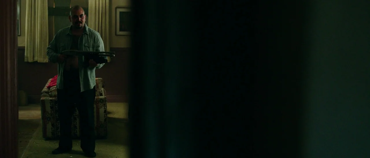 The Purge: Anarchy (2014) movie still: fullbody — A dimly lit room features a patterned armchair and beige curtains against a wall. A man stands in…