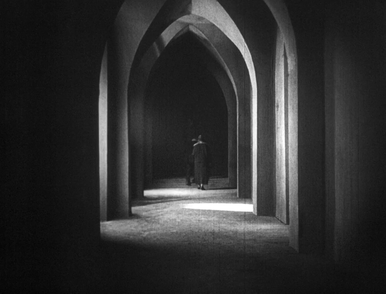 The Hands of Orlac (1924) movie still: wide — A dark stone corridor features a long row of tall, pointed Gothic arches. A lone woman walks away from…