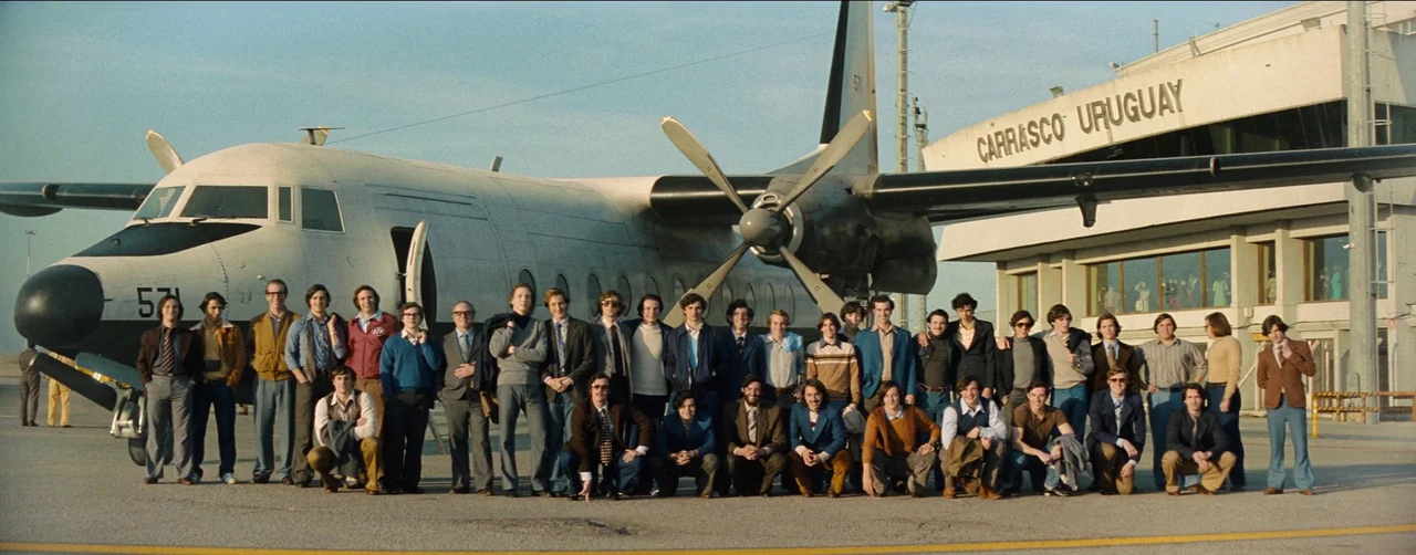 Society of the Snow (2023) movie still: wide — A large group of men in 1970s attire pose on an airport tarmac near an airplane. The airport terminal…