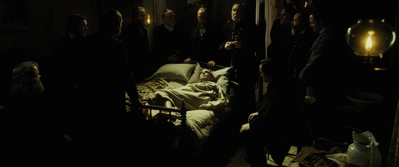 Lincoln (2012) movie still: wide — A dimly lit room in the 1860s contains a man lying on a bed. A group of formally dressed men stands gathered…