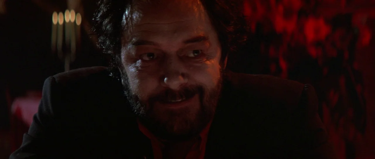 The Cook, The Thief, His Wife & Her Lover (1989) movie still: closeup — A dimly lit room is awash in deep red tones. A middle-aged man with a full…