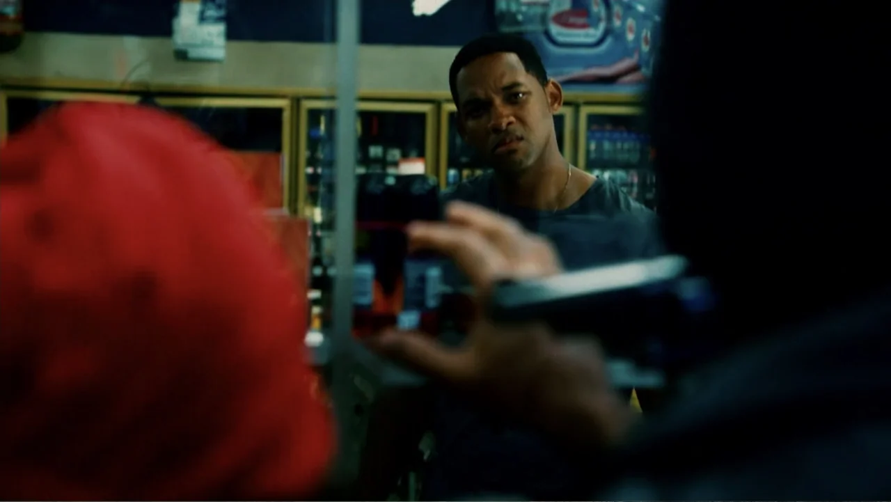 Hancock (2008) movie still: medium — A dark store interior features a glass security window separating two distinct spatial areas. A man faces…