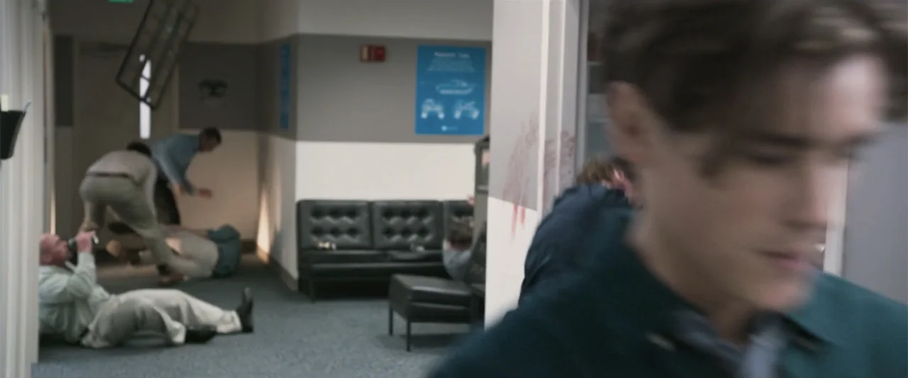Office Uprising (2018) movie still: medium — An office hallway features grey carpeting and a black button-tufted leather sofa near walls. Several…