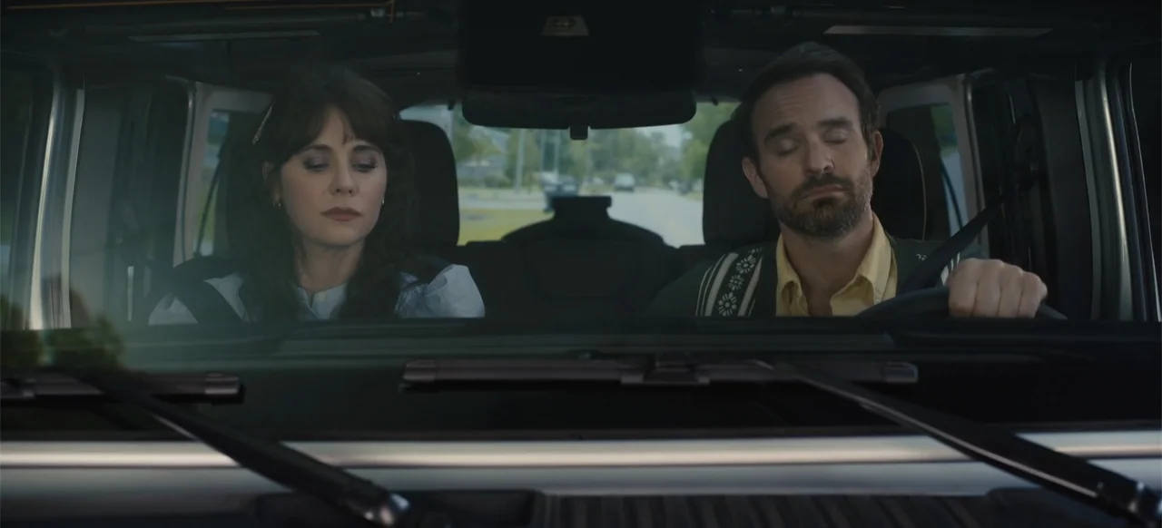 Merv (2025) movie still: medium — The interior of a modern vehicle viewed from the front passenger perspective. A man drives while a woman sits in…