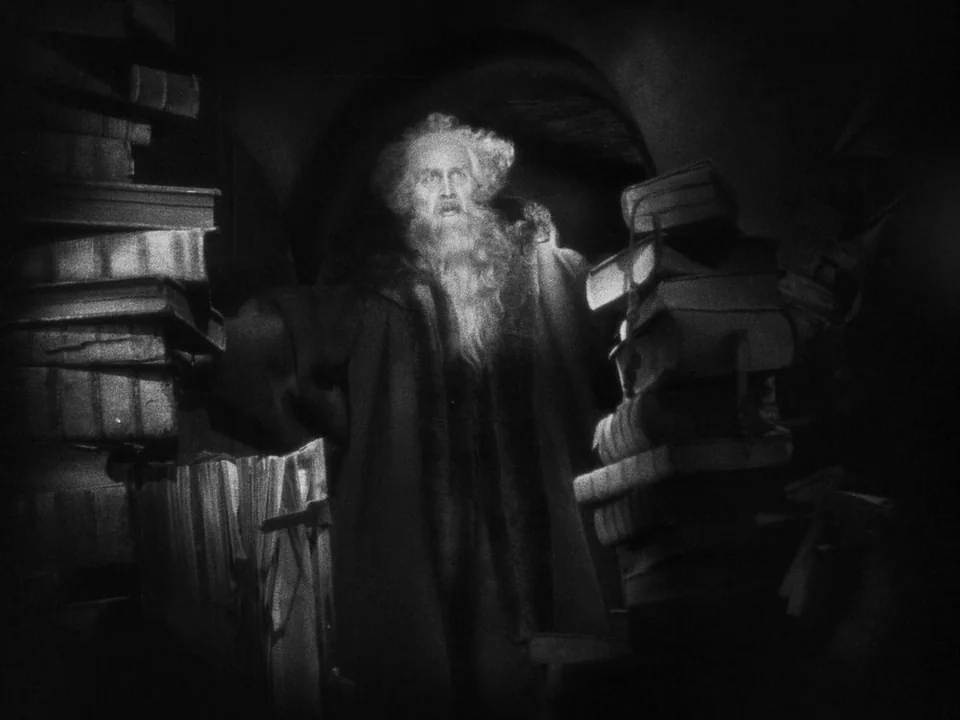 Faust (1926) movie still: medium — A dimly lit room with towering stacks of antique books flanking an elderly man. The man stands between the book…