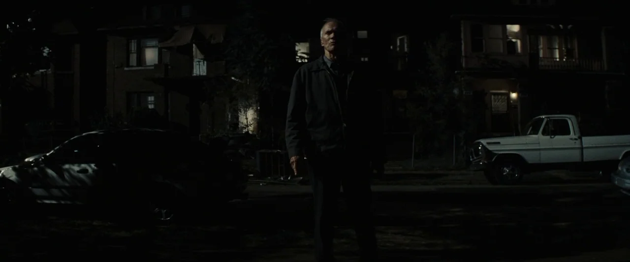 Gran Torino (2008) movie still: fullbody — An elderly man stands in the center of a dark residential street at night. Two parked vehicles flank him…