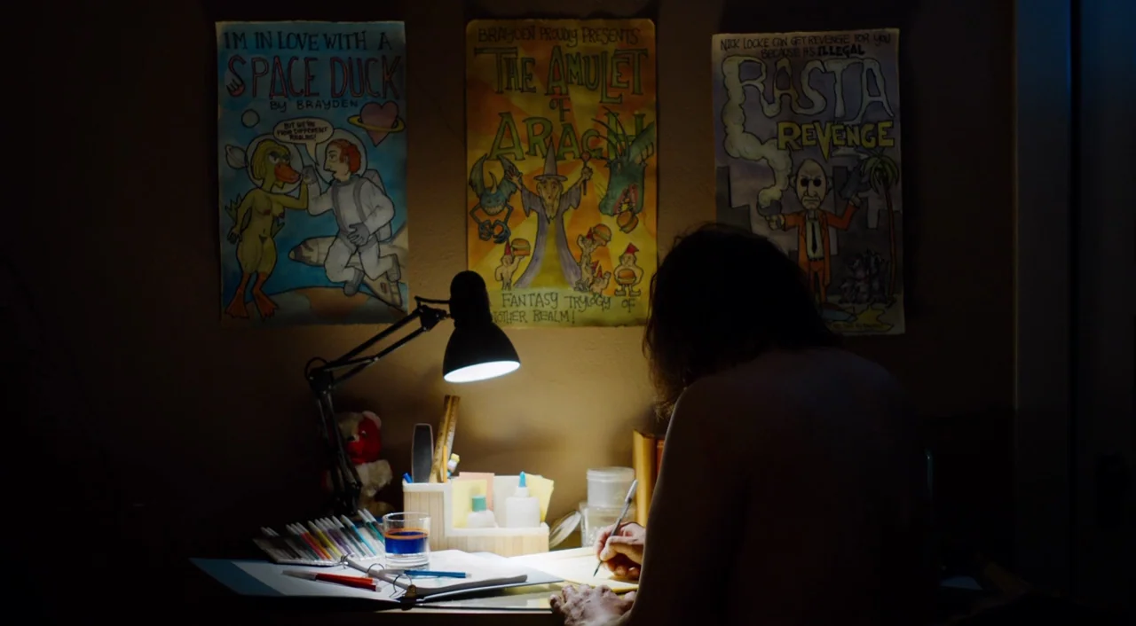The Greasy Strangler (2016) movie still: medium — A dimly lit room features a person hunched over a cluttered wooden desk. Three hand-drawn posters…