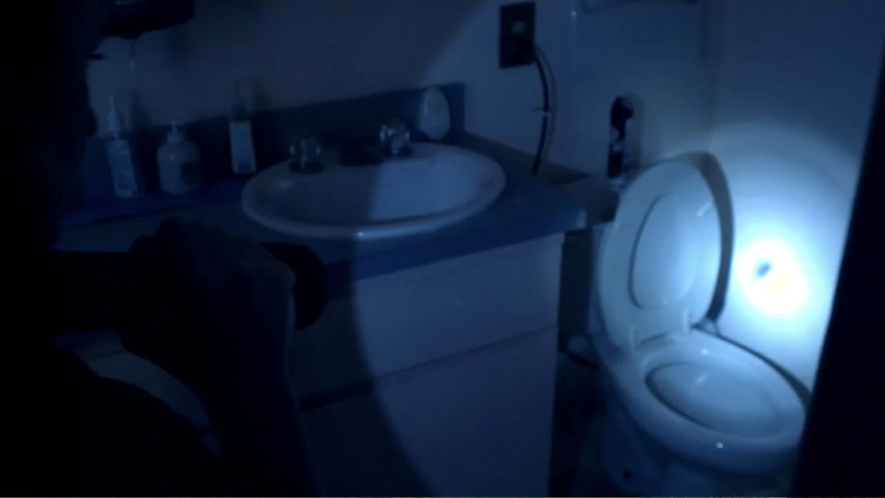 Abandoned Dead (2015) movie still: medium — A small bathroom at night contains a sink vanity and a white toilet. An unseen person aims a bright…