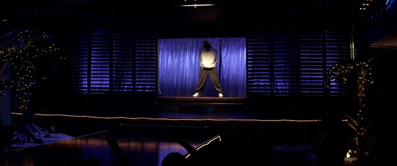 Magic Mike (2012) movie still: wide — A darkened nightclub features a center stage illuminated by a blue curtain backdrop. A person wearing a white…