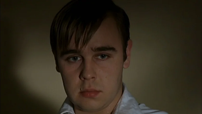 Funny Games (1997) movie still: closeup — A man stands in a dimly lit room with plain, dark walls. He looks directly toward the camera with a steady…