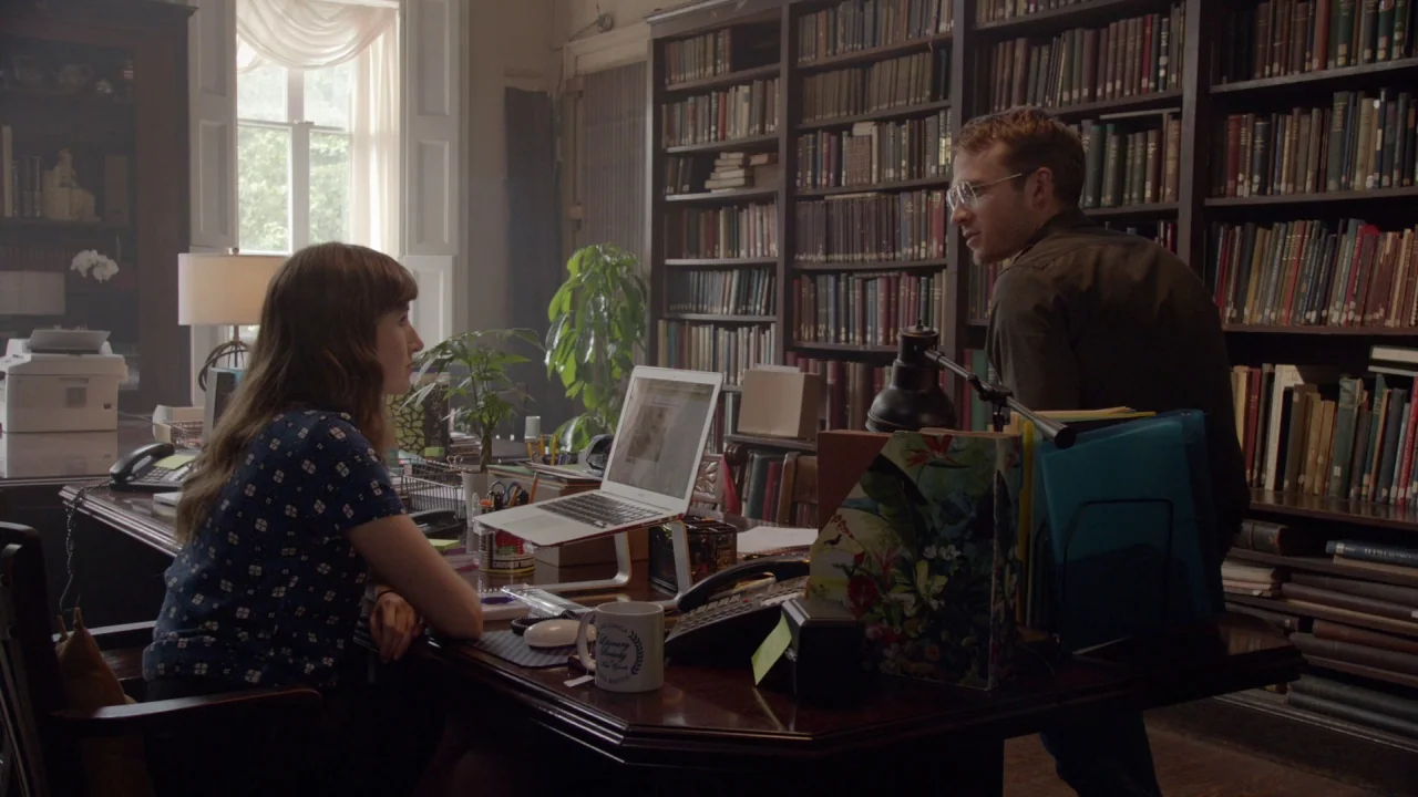 The Heart Machine (2014) movie still: medium — A cluttered room features dark wood bookshelves packed with books covering the walls. Two people…