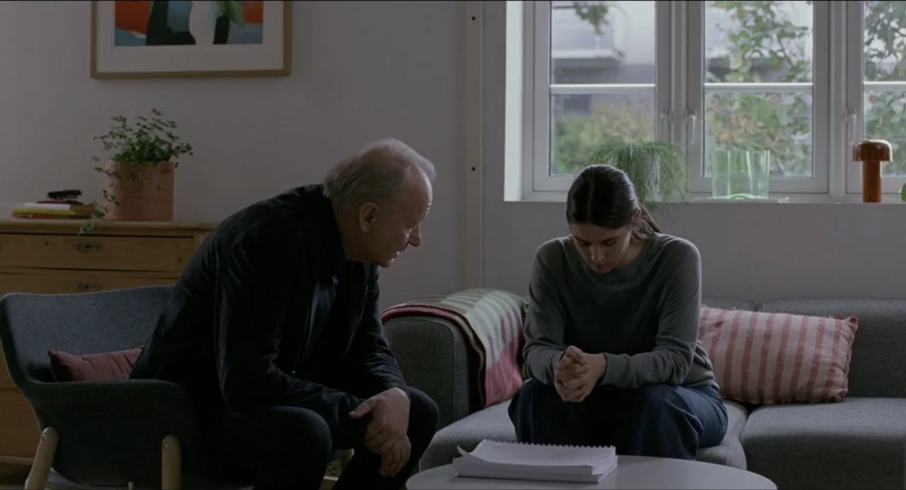 Sentimental Value (2025) movie still: medium — A quiet living room with a grey sofa and a wooden dresser near a window. An older man sits opposite a…
