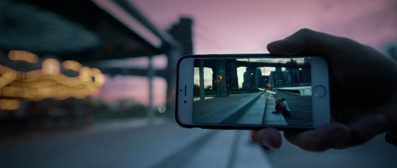 Nerve (2016) movie still: closeup — A hand holds a smartphone up to capture a view of a bridge and city skyline. The background shows a blurred…