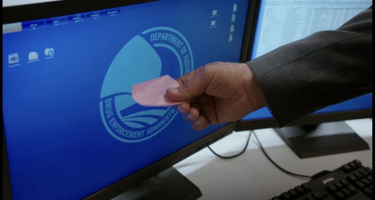 Sabotage (2014) movie still: closeup — An office workspace features dual computer monitors displaying the Department of Justice seal. A man's hand…