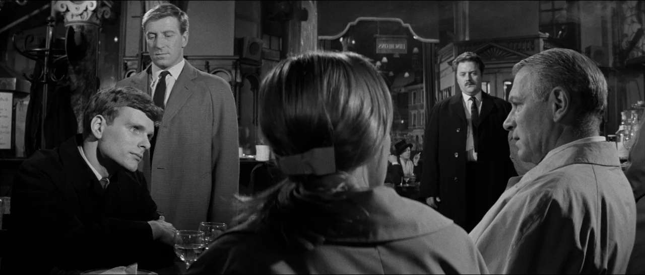 Bunny Lake Is Missing (1965) movie still: medium — A dimly lit restaurant interior with wooden tables and ornate dark fixtures. Four men and one…