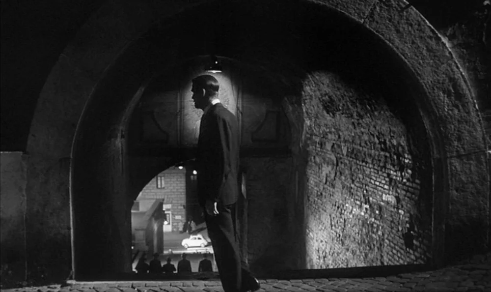 Eva (1962) movie still: fullbody — A man stands under a stone archway at night, framed by deep shadow. Below the arch, a dimly lit road with a small…