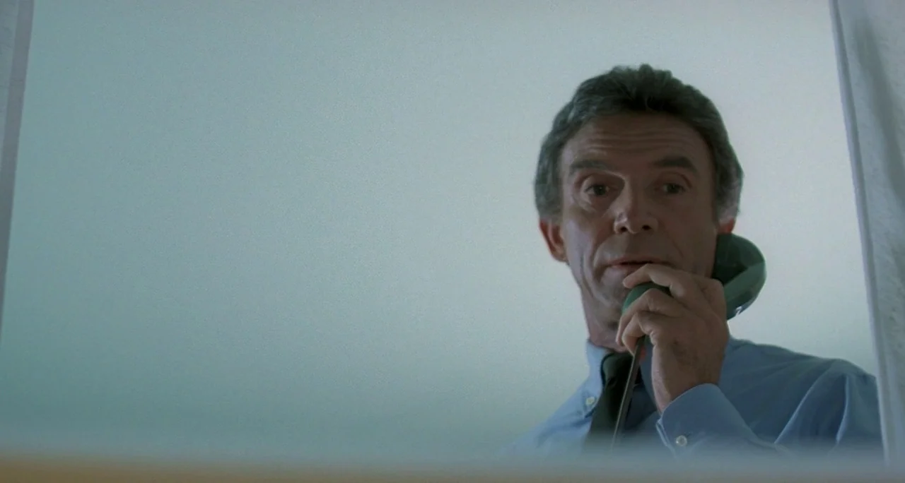 Tenebre (1982) movie still: medium — A man stands before a reflective glass surface in a sterile, brightly lit space. He holds a vintage green…