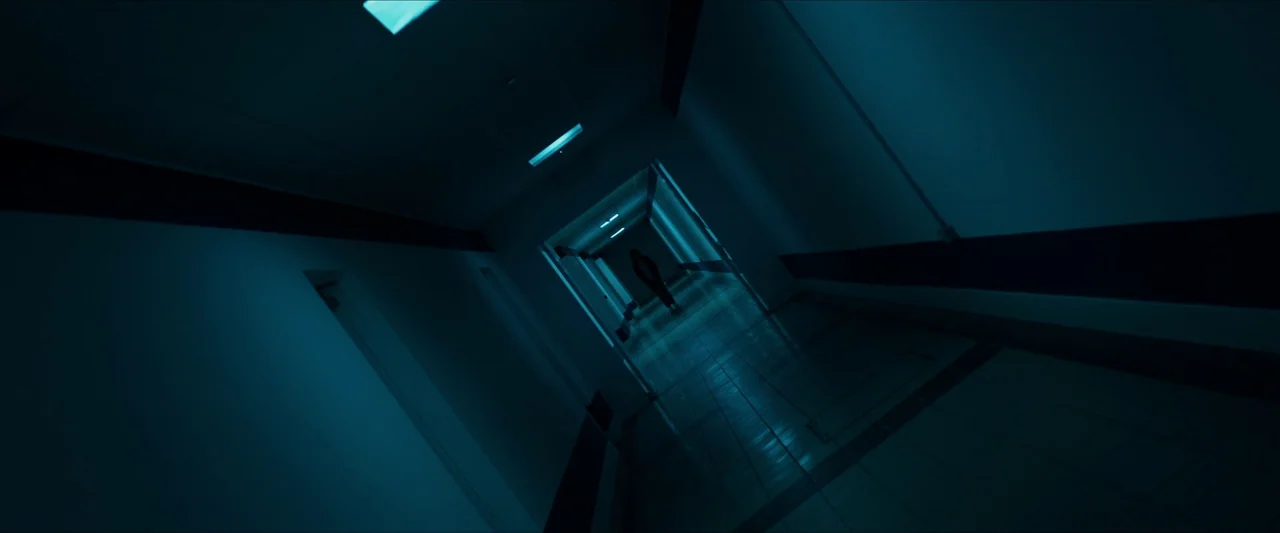 Double Blind (2023) movie still: wide — A long, hospital hallway is tilted at a severe Dutch angle with dark blue lighting. A single silhouetted…