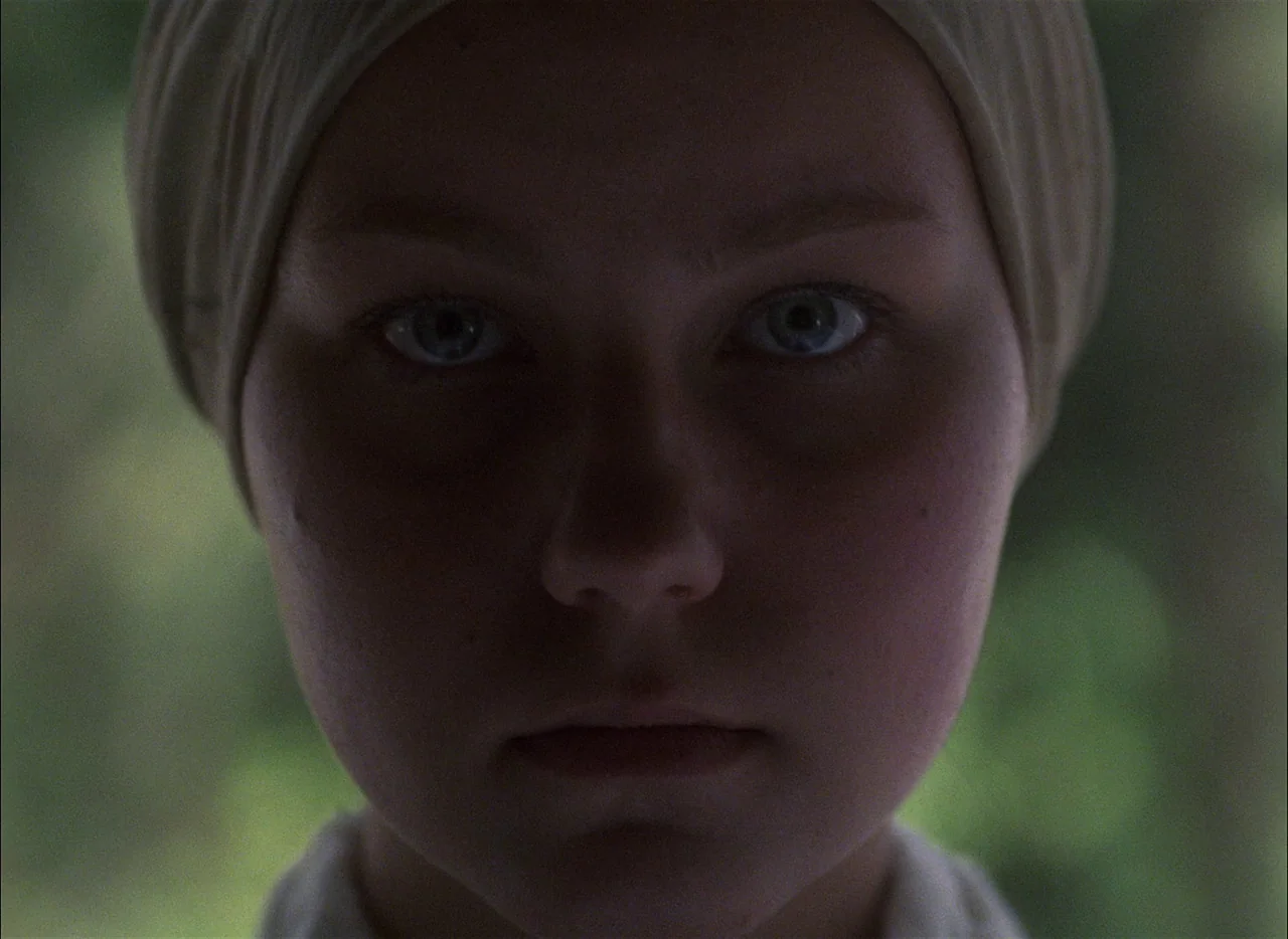 Come and See (1985) movie still: closeup — A close portrait shot of a young woman standing in a forest. She stares directly into the lens while…