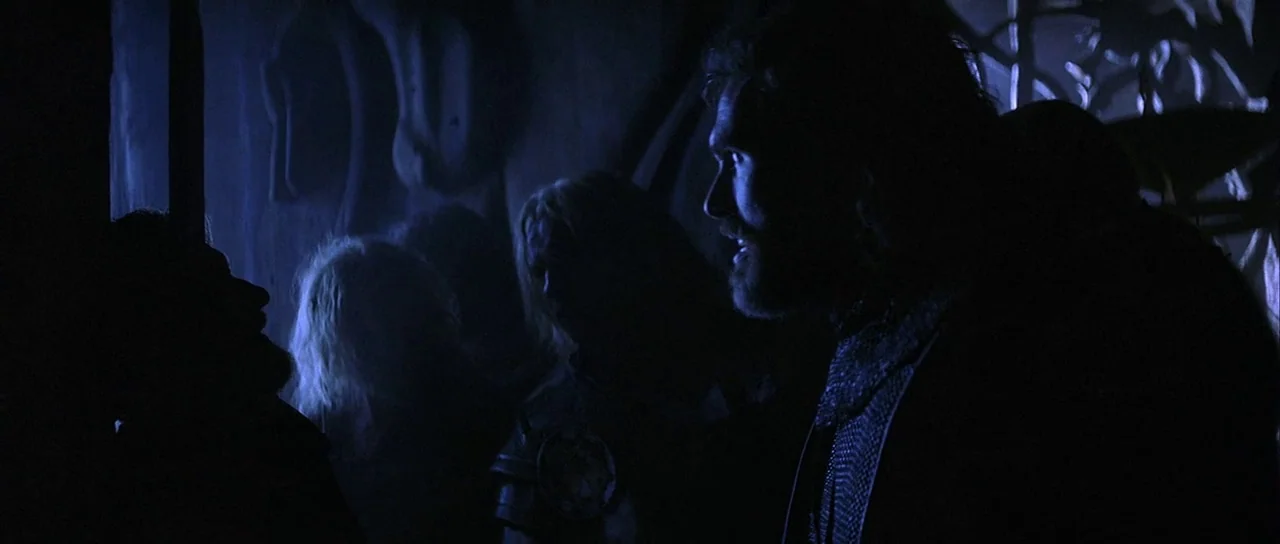 The 13th Warrior (1999) movie still: medium — A dark, stone-walled chamber features carved relief designs on the vertical masonry surface. Two men…