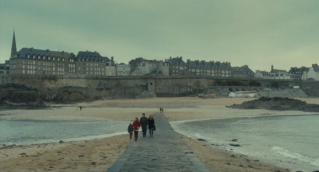 Things to Come (2016) movie still: wide — A stone causeway stretches across a beach toward a historic town with thick walls. Four people walk in a…